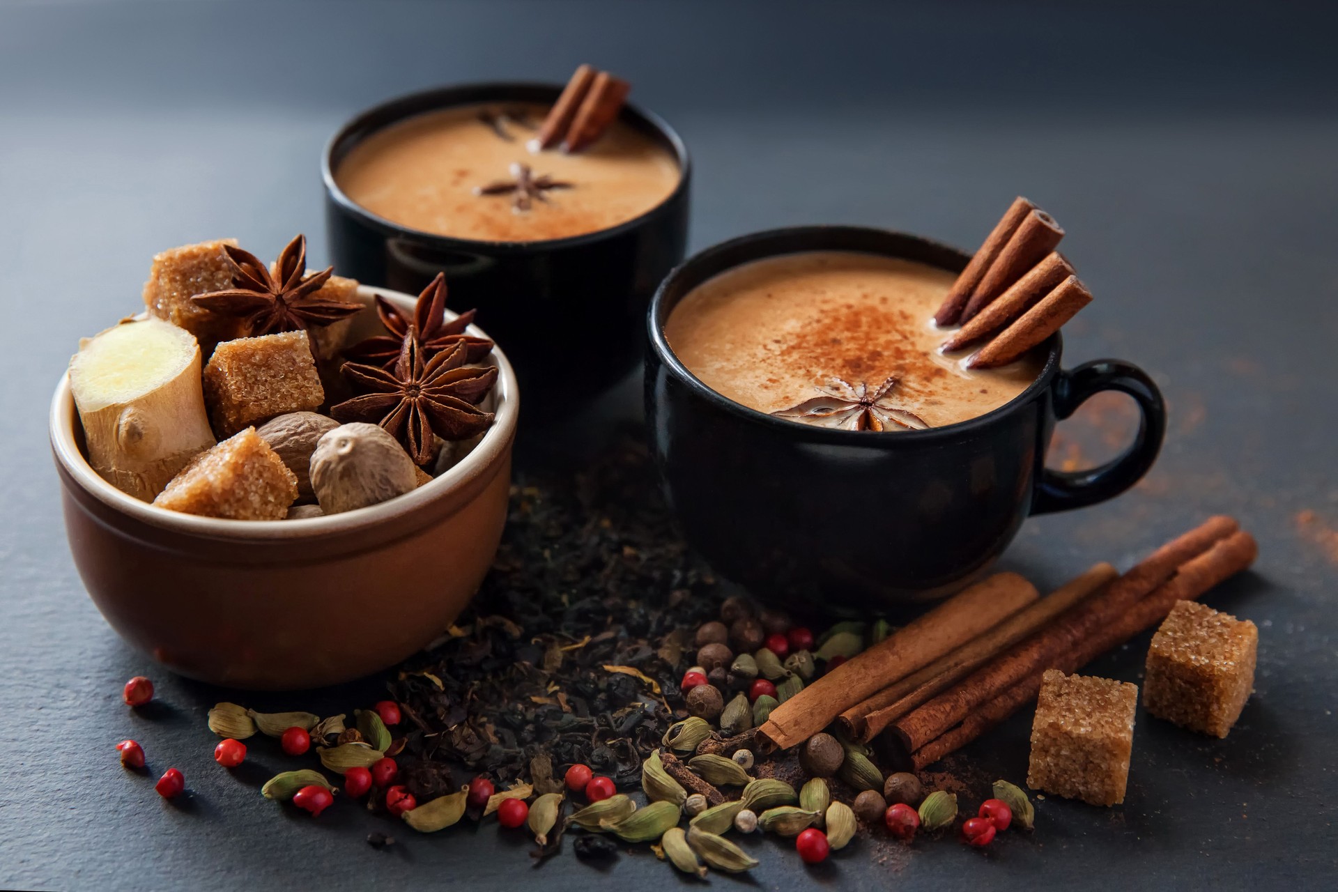Masala tea traditional hot Indian sweet milk spiced drink in black cups. Spices cinnamon sticks, cardamom pods, star anise, sugar, nutmeg, coriander and pepper seeds in brown ceramic cup on a black slate stone plate